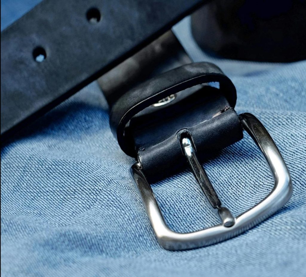 Close-up of a black leather belt with metal buckle laid on blue denim fabric.